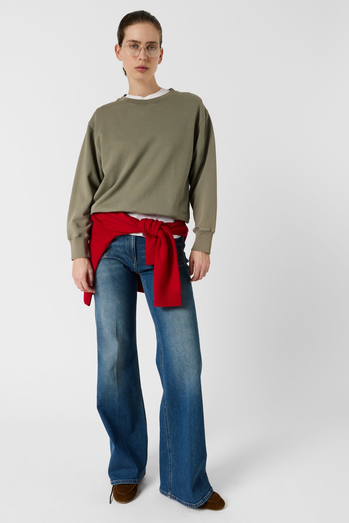 A person wearing glasses, an olive sweatshirt, flared blue jeans, brown shoes, and a red sweater tied around their waist stands against a plain white background.