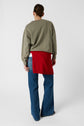 A person stands facing away, wearing a greenish-gray sweater, a red sweater tied around their waist, blue jeans, and beige shoes, with their hair pulled back. The background is plain white.