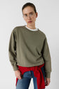 A woman with glasses wears an olive green sweatshirt, white shirt, and blue jeans. A red sweater is tied around her waist. She stands against a plain white background, looking directly at the camera.