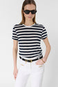 A person wearing black sunglasses, a black and white striped t-shirt, white pants with gold accents, and a black belt, stands against a plain white background.