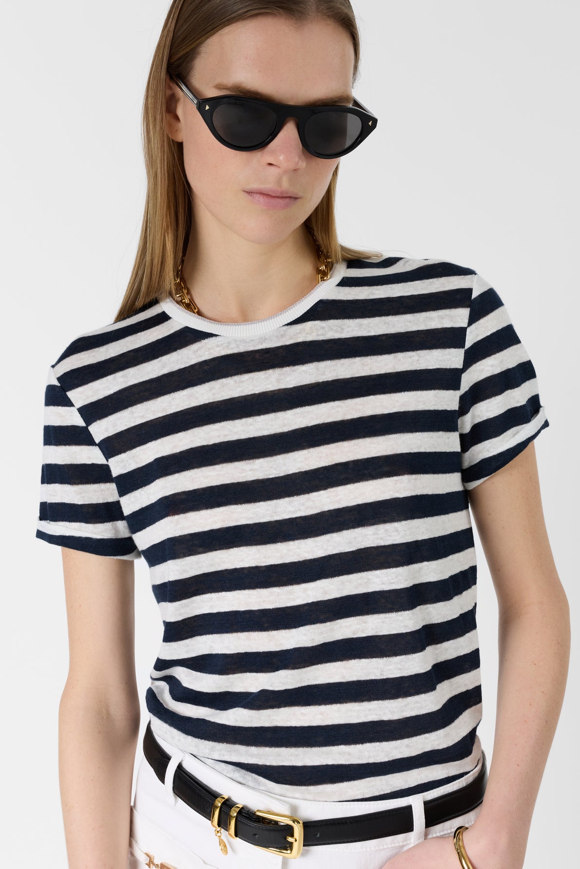 Woman wearing black sunglasses and a black-and-white striped t-shirt, with white pants and a black belt with a gold buckle, standing against a plain background.