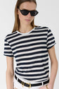 A person wearing black sunglasses and a navy-and-white striped t-shirt stands with one hand in a pocket. They have straight hair and are also wearing a black belt with a gold buckle.