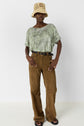 A person wears a tan bucket hat, a green snakeskin-patterned top, brown belted pants, and black sandals. They stand against a plain white wall, posing with one hand in their pocket.