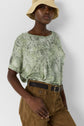Woman wearing a light green patterned top, tan wide-brim bucket hat, brown high-waisted pants with a black belt, standing against a plain white background.