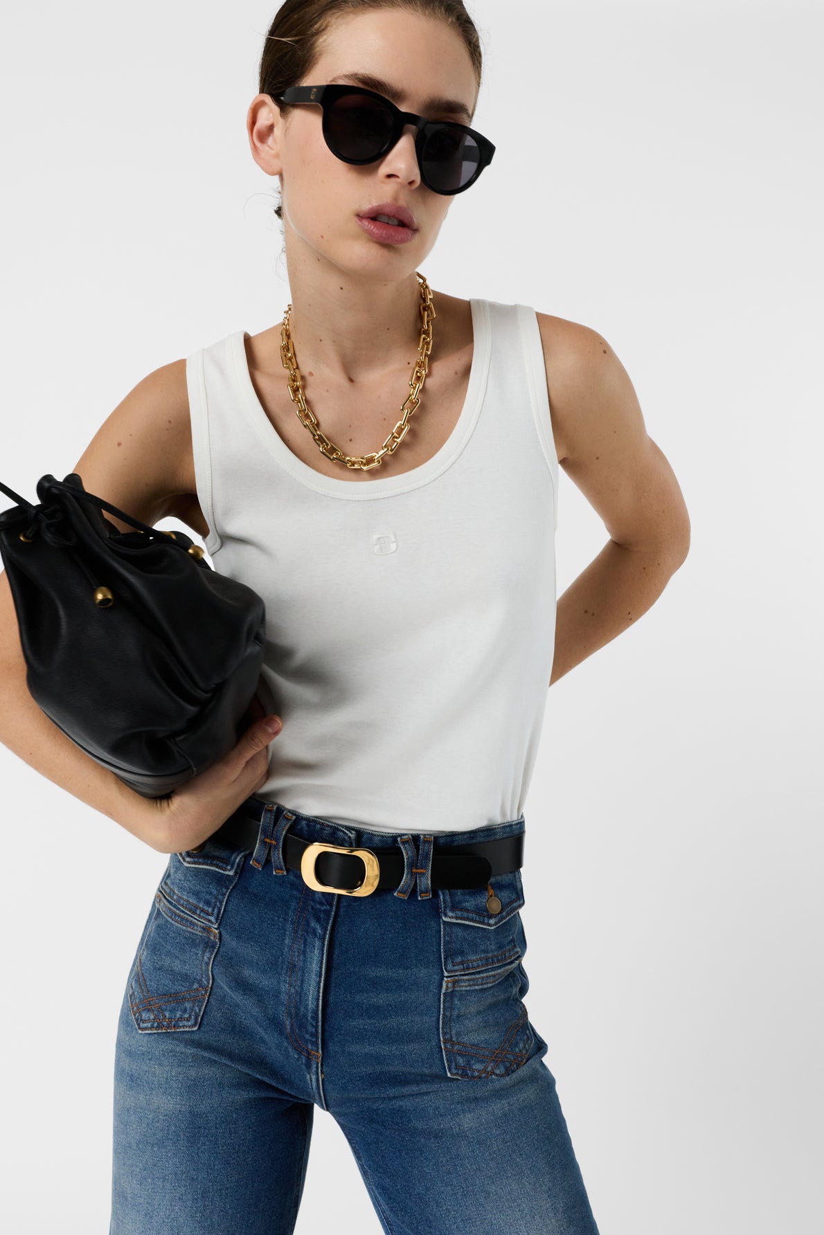Woman wearing sunglasses, a débardeur blanc en coton with a monogramme brodé, gold chain necklace, and high-waisted blue jeans holds a black bag under her arm against a plain white background.