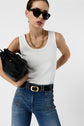 Woman wearing sunglasses, a débardeur blanc en coton with a monogramme brodé, gold chain necklace, and high-waisted blue jeans holds a black bag under her arm against a plain white background.
