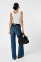 Woman standing with her back to the camera, wearing a débardeur blanc en coton, blue jeans, black belt, and black shoes. She holds a large black handbag in her left hand and has brown hair pulled back. The top features a delicate monogramme brodé, adding an elegant touch to her outfit.