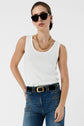 Woman wearing black sunglasses, a gold chain necklace, and a débardeur blanc en coton with monogramme brodé, paired with high-waisted blue jeans and a black belt, posing with one hand on her hip against a plain background.