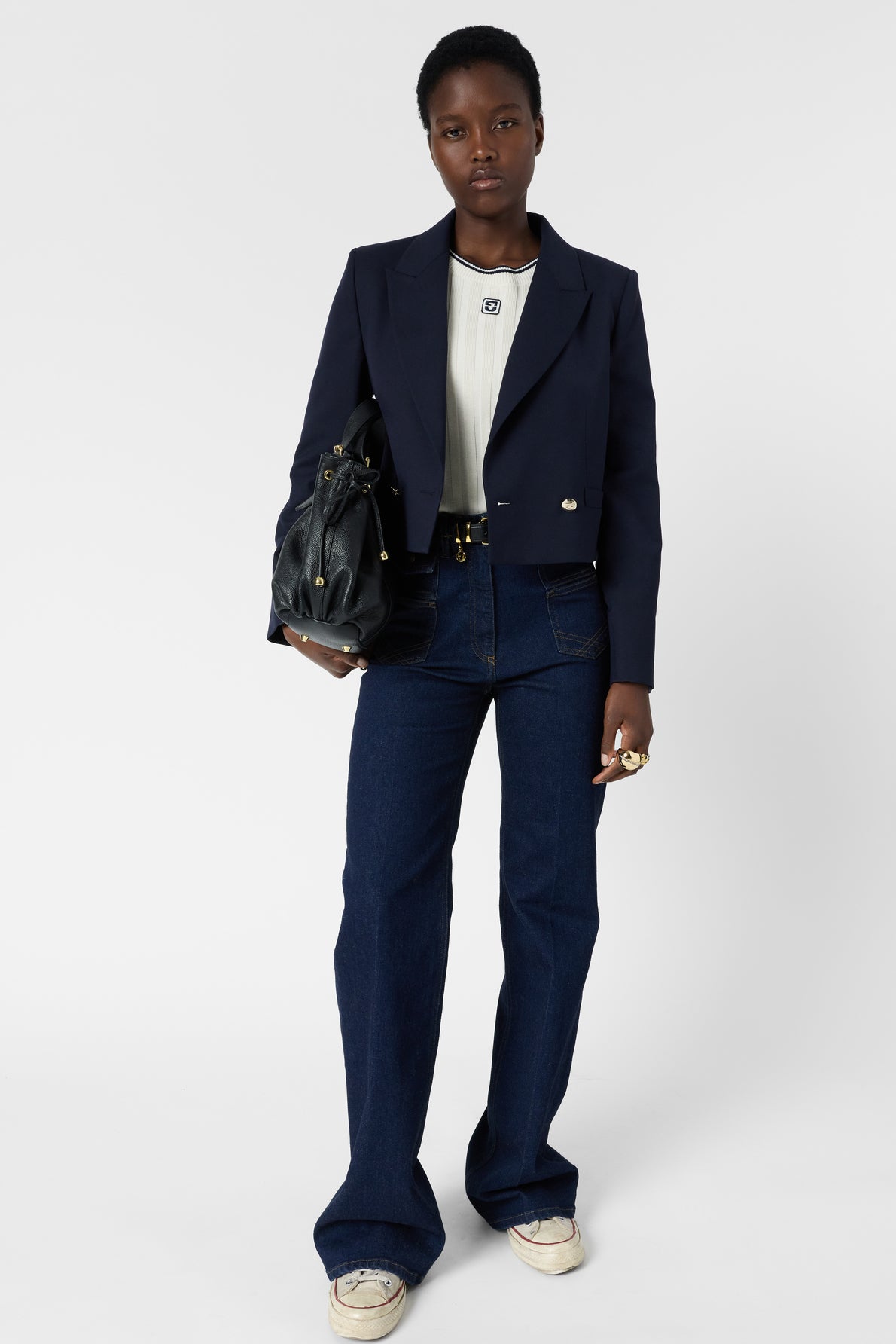 A person stands against a white background wearing a navy blazer, white shirt, blue jeans, white sneakers, and holding a black bag. They have short hair and wear rings on their fingers.