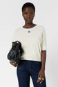 A person wearing a white ribbed short-sleeve sweater and dark jeans holds a black handbag with gold accents, standing against a plain white background.
