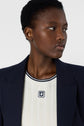 A person with short curly hair wears a navy blazer over a white ribbed shirt with a navy logo, looking to the side against a plain light background.