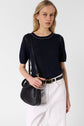 A woman wearing a navy short-sleeve top, white pants, and a black belt stands against a plain background, holding a black shoulder bag.