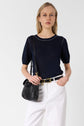 Woman with long blonde hair wears a navy short-sleeve top, white pants, and a black belt. She carries a small black shoulder bag, standing against a plain light background.
