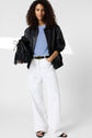 Woman with curly hair wears a black leather jacket over a light blue shirt, white wide-leg pants, and black sandals, holding a black fringed handbag. She stands confidently against a plain light background.