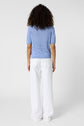 A person with curly hair, wearing a light blue short-sleeve sweater and white wide-leg pants, stands facing away against a plain white background.