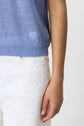 A person wearing a light blue shirt with a small embroidered logo and white pants, shown from the waist to mid-arm. Their right arm is relaxed at their side against a plain background.
