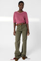A person stands wearing a pink short-sleeve top, green patterned high-waisted pants, a brown woven belt, and brown shoes with tassels, posing against a plain white background.