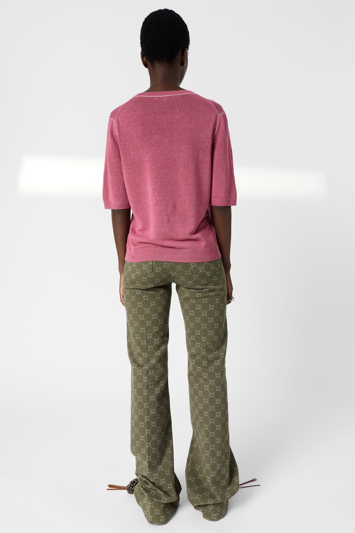 A person stands facing away, wearing a pink short-sleeve shirt and olive green patterned pants with flared legs. The background is plain white.