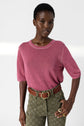 A person wearing a pink short-sleeve top and olive green patterned pants with a braided brown belt poses confidently against a plain white background.