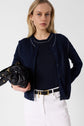 A woman with long blonde hair wears a bleu marine gilet col rond over a matching top, white pants, and a black belt. She holds a black handbag and stands against a plain white background.