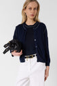A woman with long blonde hair wears a bleu marine gilet col rond over a matching top in lin mélangé, paired with white pants and holding a black leather handbag. She stands against a plain light background.
