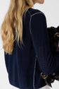 A woman with long blonde hair, wearing a bleu marine gilet col rond in a soft lin mélangé, holds a black bag with gold hardware. She is turned away from the camera.