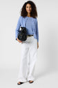 Woman with curly hair wearing a gilet bleu ciel over a blue shirt, white wide-leg pants, and black sandals. She holds a black handbag and stands against a plain white background.