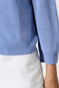 A person wearing a light blue knit sweater, styled as a gilet bleu ciel with a subtle embroidered logo near the waist and white pants. The image shows part of their torso and arm.