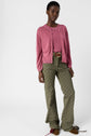 A person wearing a pink gilet femme with a col rond over a matching top, olive green patterned pants with a brown belt, and brown shoes stands against a plain white background.