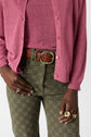 A person wearing a pink top, a pink gilet col rond, olive green patterned pants, a brown braided belt, and several gold and silver rings on their fingers.