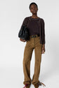 A person stands facing forward, wearing a brown cardigan, matching top, tan suede pants, and brown shoes. They hold a black textured bag and wear a belt. The background is plain white.