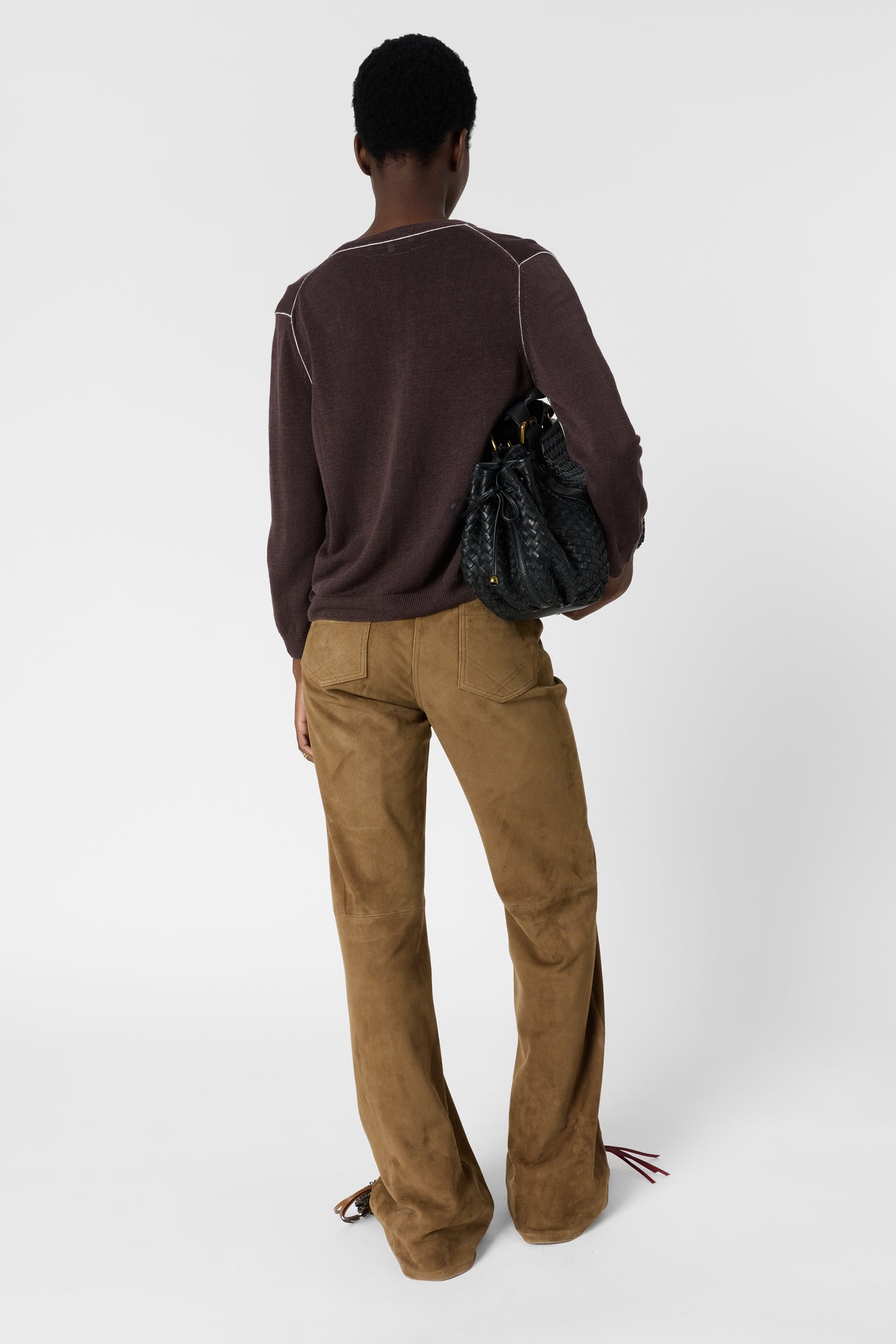 Person standing with back to camera, wearing a brown long-sleeve top and tan suede pants, holding a black textured handbag in their left arm against a plain white background.