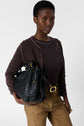 A person wearing a brown sweater and tan pants holds a large black woven handbag with gold accents, standing against a plain light background.