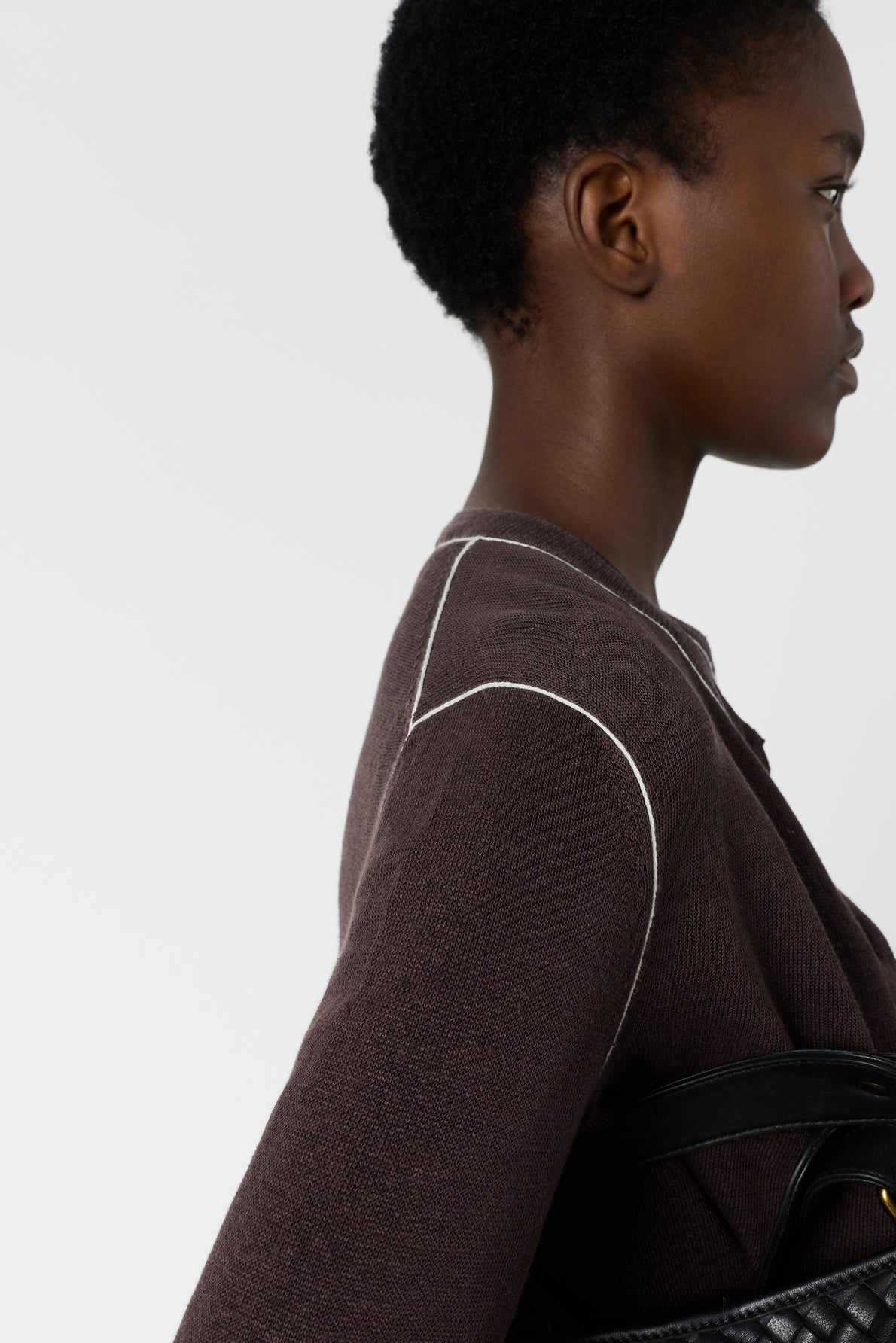A person with short curly hair, wearing a brown textured top with white stitching, stands in profile holding a black quilted bag under their arm against a plain light background.