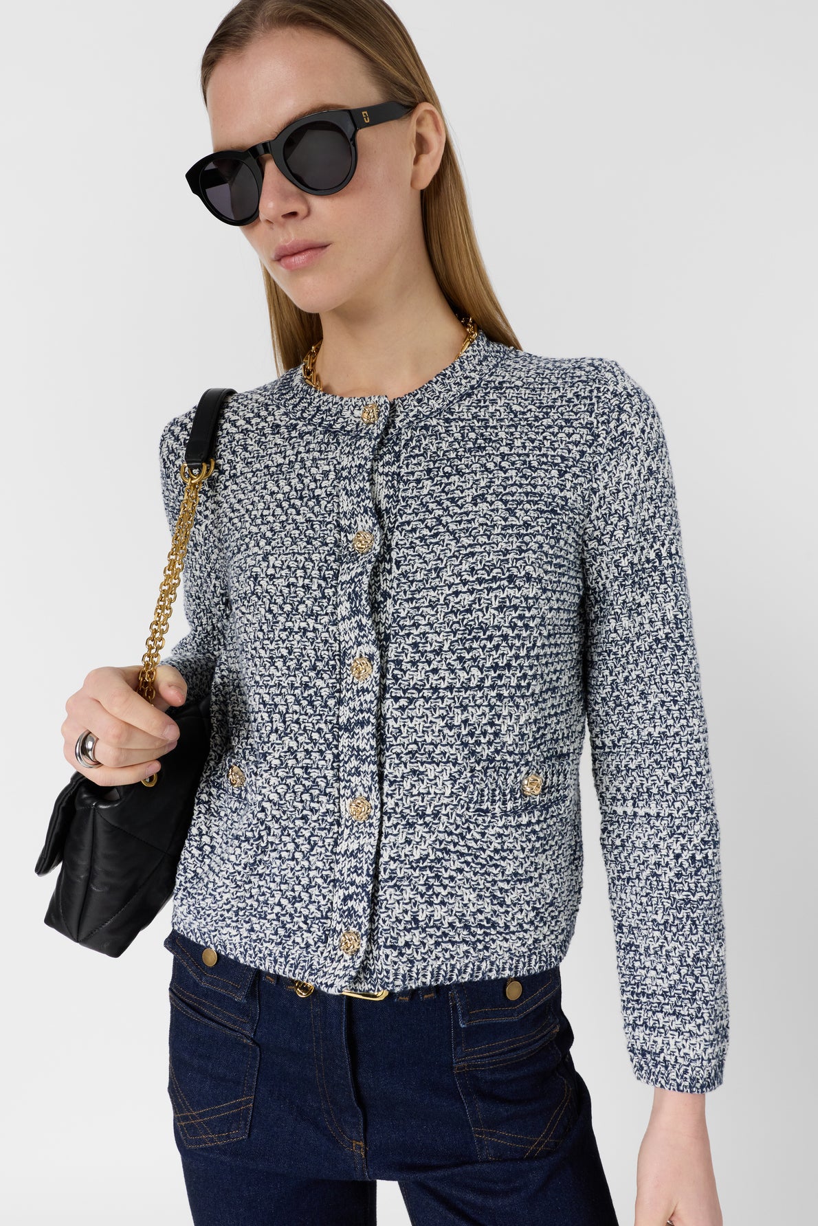 Woman wearing black sunglasses, a gilet bleu blanc à boutons dorés with a textured finish, dark jeans, and holding a black purse with a gold chain strap. She stands against a plain light background.