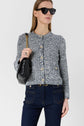 Woman wearing sunglasses, a textured gray gilet en coton chiné with gold buttons, dark blue jeans, and carrying a black quilted shoulder bag with a gold chain, standing against a plain light background.