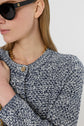 A woman wearing black sunglasses and a gilet bleu blanc—a blue-and-white textured knit top—paired with a gold chain necklace, shown from the shoulders up, facing left. Her hand is near her chest, holding the chain.