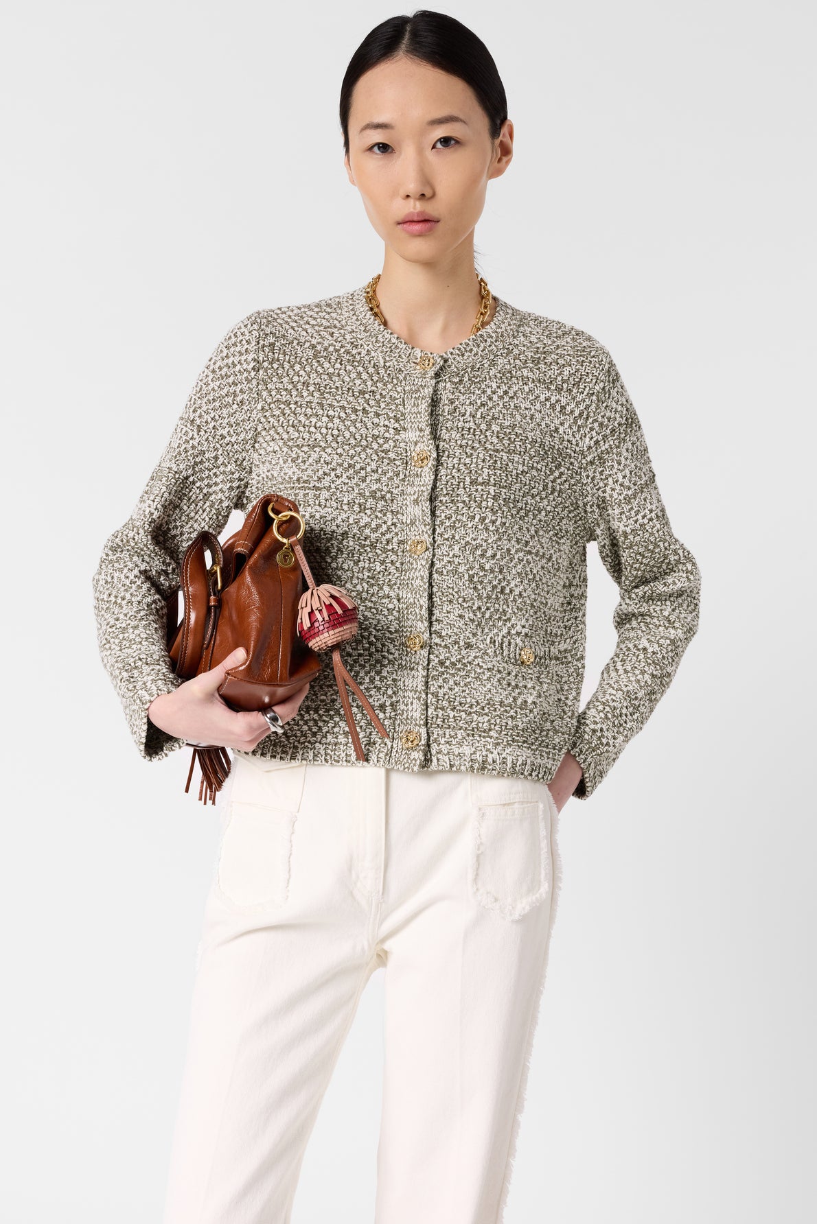 Woman wearing a textured gray gilet coton chiné and white pants holds a brown leather handbag with a gold chain strap and red charm against a plain light background.