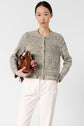 Woman wearing a textured gray gilet coton chiné and white pants holds a brown leather handbag with a gold chain strap and red charm against a plain light background.