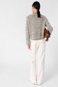 A person with long dark hair in a low ponytail, wearing a textured gray gilet coton chiné sweater and loose white pants, stands facing away, holding a brown handbag on their left shoulder against a plain white background.
