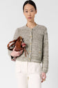 A woman with straight dark hair wears a gilet court femme in textured gray cotton, styled as a gilet coton chiné, paired with white pants and holding a brown handbag with keychains against a plain light background.