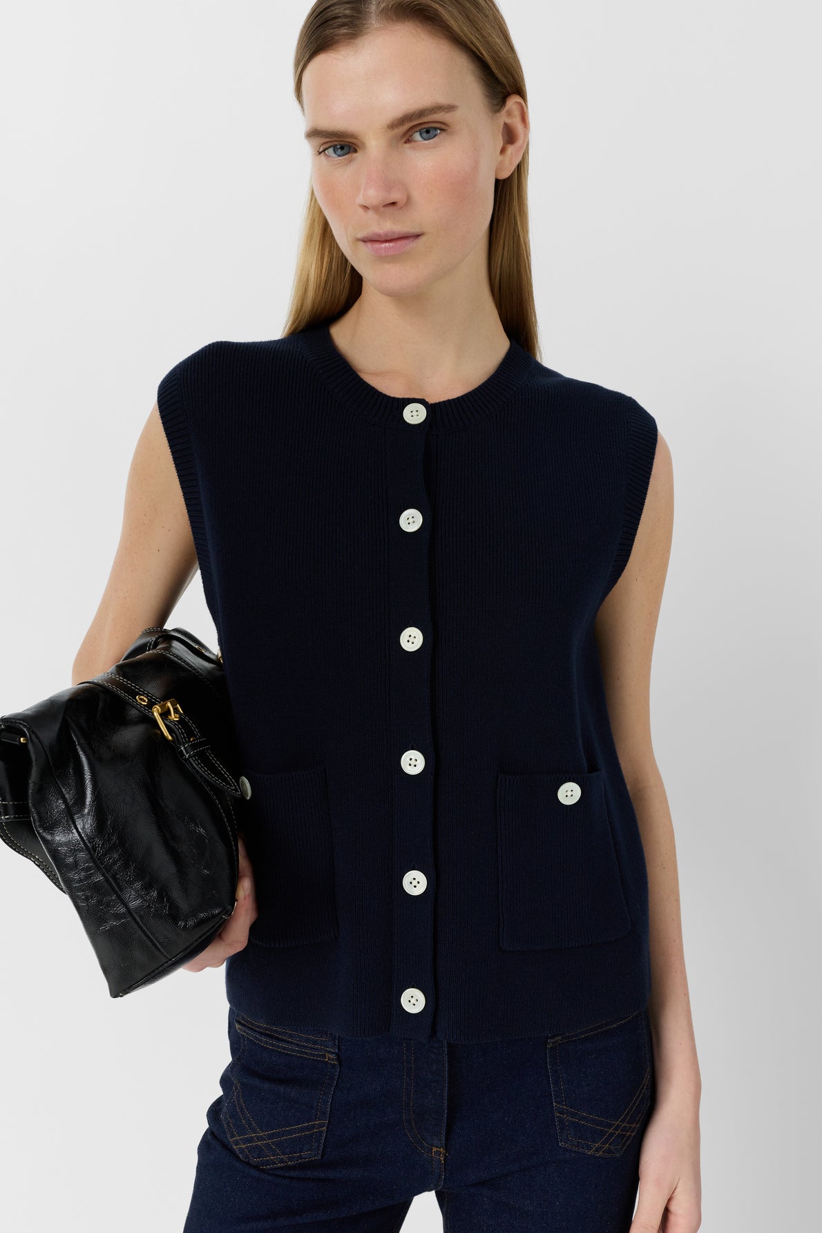 A person wearing a gilet boutonné sans manche en coton bleu marine with front pockets and dark jeans holds a black leather bag.