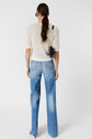 A person with long brown hair in a ponytail wears a white knit short-sleeve top, light blue jeans, black heels, and carries a small black handbag, standing against a plain white background, facing away.