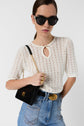 A woman wearing black sunglasses, a white knit short-sleeve top, blue high-waisted jeans with a gold buckle belt, and holding a black purse with a gold chain strap over her shoulder.