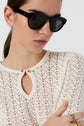 Woman wearing black sunglasses, a white lace top with a keyhole neckline and gold buttons, and gold rings, posing against a plain background.