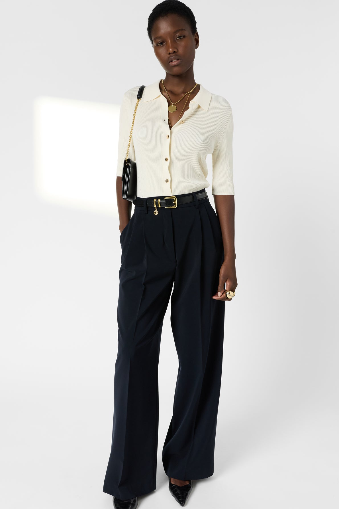A person stands against a white background wearing a white maille côtelée gilet polo with boutons dorés, black wide-leg trousers, black heels, and gold jewelry, with a black shoulder bag. Hands are in pockets, and gaze is directed forward.