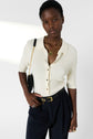 A person wearing a cream maille côtelée ribbed button-up top with boutons dorés, dark pleated pants, layered gold necklaces, and a black belt poses with a black shoulder bag against a plain, softly lit background.