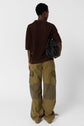 Person standing with back to camera, wearing a dark brown short-sleeve sweater and loose olive green cargo pants, holding a textured black handbag on one shoulder against a plain white background.