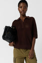 A person wearing a dark brown short-sleeve knit top and olive green pants holds a black woven handbag against a plain light background.