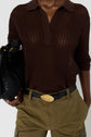A person wearing a brown crocheted top, olive green pants with a large belt buckle, and holding a black woven handbag.